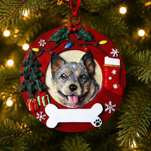 A round red Christmas ornament hanging on a lit Christmas tree. The ornament features a circular cutout with an Australian Cattledog in the center, surrounded by holiday decorations including a small tree, stockings, snowflakes, a bone, and colorful lights.