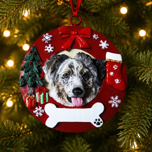 A personalized Christmas ornament shaped like a red wreath hanging on a pine tree. The ornament features a custom pet portrait of an Australian Shepherd, surrounded by holiday decorations including a red bow, white snowflakes, a small Christmas tree, a red stocking, a white bone, and gift boxes.