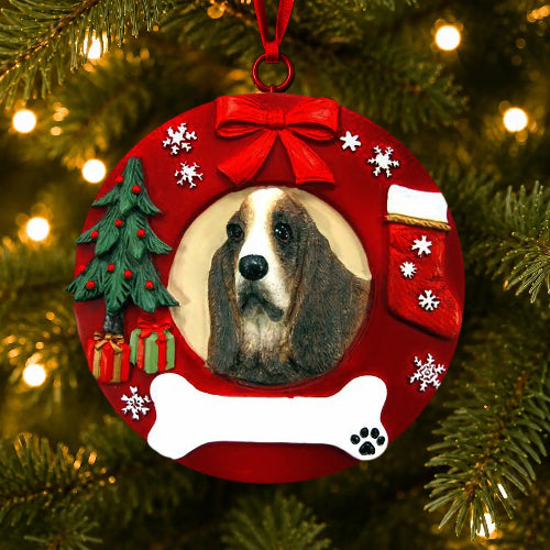 A round red Christmas ornament hanging on a Christmas tree, featuring a photo cutout of a Basset Hound, a red bow, a small tree, gifts, snowflakes, a stocking, and a white bone with a blank paw-print spot.