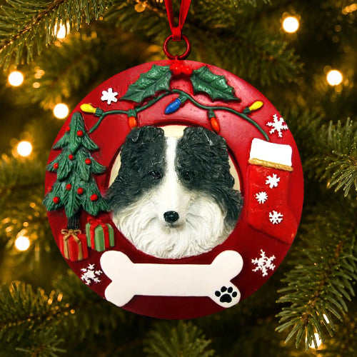 A round red Christmas ornament hanging on a Christmas tree, featuring a custom pet portrait of a Border Collie surrounded by holiday decorations like a tree, stocking, bone, and ornaments.