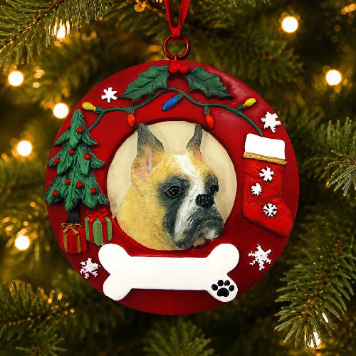 A round red Christmas ornament hanging on a lit Christmas tree. The ornament features a raised red border with holiday decorations including a green tree, stockings, bone, and lily of the valley, with a circular inset showing a  Boxer with cropped ears dog portrait.