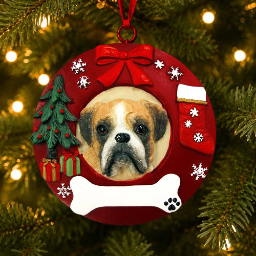 A round red Christmas ornament shaped like a wreath, hanging on a lit Christmas tree. The ornament features a custom photo cutout of a Boxer with uncropped ears , surrounded by festive decorations including a red bow, white snowflakes, a small green Christmas tree, a red stocking, and a white bone at the bottom.