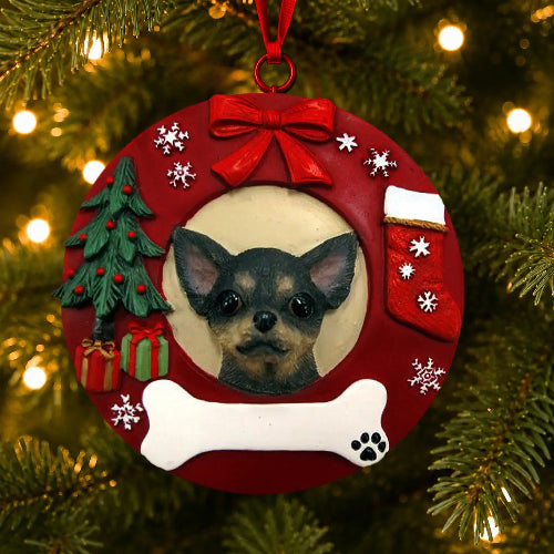 A circular red Christmas ornament hanging on a Christmas tree, featuring a portrait of a Black Chihuahua dog in the center, surrounded by a red bow, white snowflakes, a small green tree, a red stocking, a white bone, and a gift box.