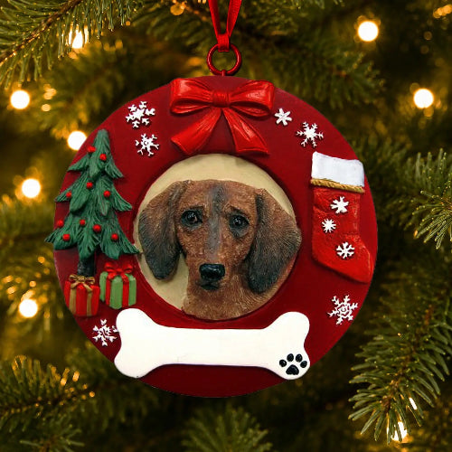 A circular red Christmas ornament shaped like a wreath, featuring a portrait of a Red Dachshund dog in the center, a red bow at the top, a white bone at the bottom, a small green Christmas tree, a red stocking, and snowflake decorations, hanging on a Christmas tree with warm lights.