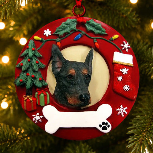 A circular red Christmas ornament shaped like a wreath, hanging on a Christmas tree. The ornament features a custom pet portrait of a black and tan Doberman dog in the center, surrounded by holiday decorations including a green Christmas tree, red ornaments, a red stocking, a white bone, and a white paw print detail.