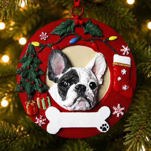 A circular red Christmas ornament featuring a raised black-and-white French Bulldog portrait, hanging on a Christmas tree with warm lights in the background. The ornament includes holiday decorations such as a small green tree, red gift boxes, a white bone, a red stocking, and snowflake accents.