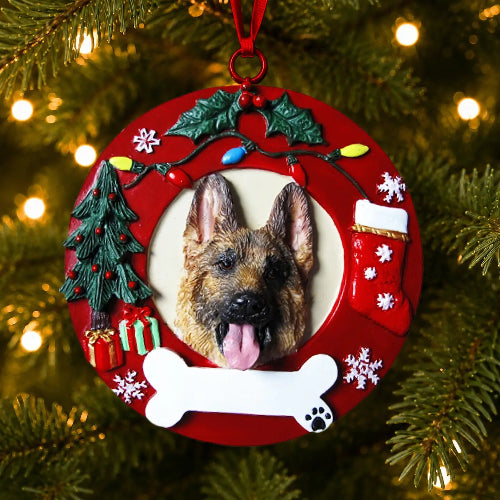 A round red Christmas ornament featuring a custom photo of a German Shepherd dog's face, surrounded by festive holiday decorations including a miniature Christmas tree, stockings, bone, holly, and lights, hanging against a blurred Christmas tree background with warm lights.
