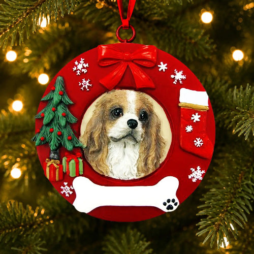 A round red Christmas ornament hanging on a Christmas tree, featuring a portrait of a Cavalier King Charles Spaniel dog, a red bow, a small green Christmas tree, a stocking, snowflakes, and a white bone at the bottom.