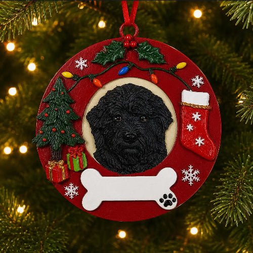 A round red Christmas ornament featuring a Black Labradoodle portrait in the center, surrounded by holiday decorations including a Christmas tree, stockings, snowflakes, lights, and a white bone, hanging on a Christmas tree with warm lights.