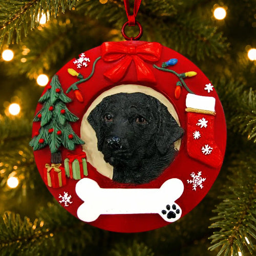 A red Christmas wreath-shaped pet ornament hanging on a lit Christmas tree. The ornament features a Black Labrador portrait in the center, a red bow at the top, a small green tree, gift boxes, colorful holiday lights, white snowflakes, a white bone shape at the bottom, and a red stocking on the right.
