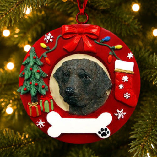 A circular red Christmas ornament hanging on a Christmas tree, featuring a portrait of a Chocolate Labrador Retriever in the center, a red bow at the top, a small green Christmas tree, gifts, a stocking, snowflakes, and a white bone at the bottom.