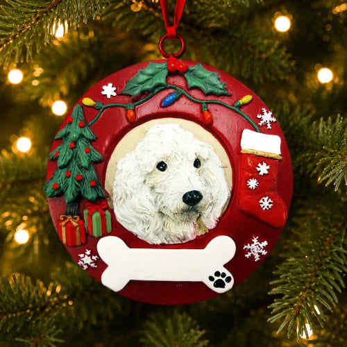 A round red Christmas ornament hanging on a pine tree, featuring a white poodle in the center surrounded by holiday decorations including a small tree, gift boxes, a stocking, snowflakes, and a bone at the bottom.