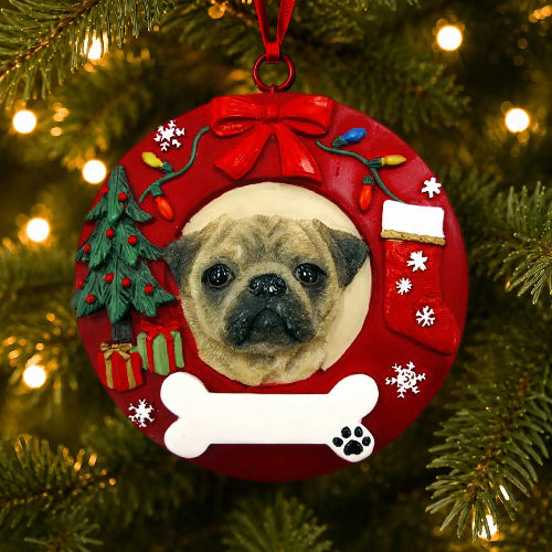 A custom round Christmas ornament featuring a portrait of a fawn pug dog, decorated with a red wreath design, a red bow, a miniature Christmas tree, a red stocking, a white bone, and snowflakes, hanging on a lit Christmas tree.