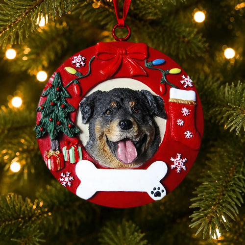 A round Christmas ornament hanging on a Christmas tree, featuring a custom pet portrait of a Rottweiler dog inside a red wreath design with a tree, gifts, snowflakes, and a white bone at the bottom.