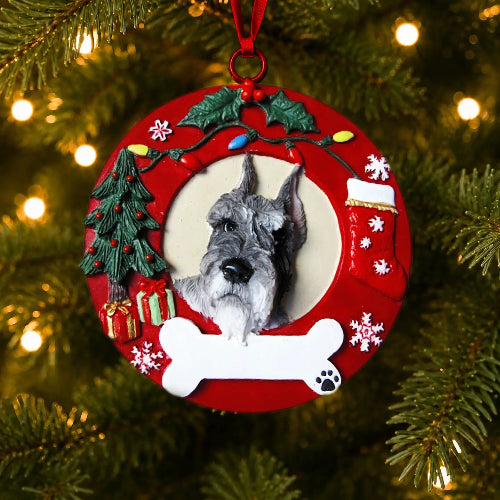A round red Christmas ornament hanging on a Christmas tree, featuring a sculpted Schnauzer with cropped ears in the center, surrounded by holiday decorations including a green tree, white bone, red stocking, and snowflakes.