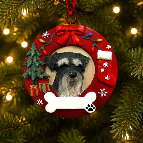 A round red Christmas ornament hanging on a lit Christmas tree. The ornament features a portrait of a Schnauzer with uncropped ears in the center, surrounded by holiday decorations including a red bow, Christmas tree, stockings, gifts, snowflakes, and a white bone at the bottom.