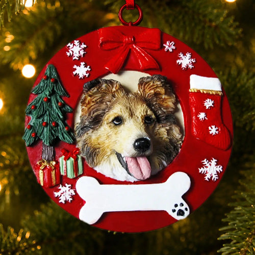A round red Christmas ornament hanging on a Christmas tree, featuring a custom photo cutout of a Sheltie dog's face, surrounded by festive decorations including a red bow, white snowflakes, a small green Christmas tree, wrapped gifts, a red stocking, and a white bone-shaped cutout.
