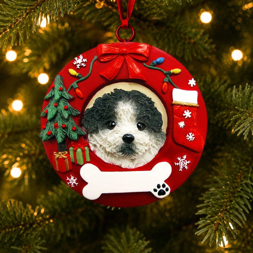 A round red Christmas ornament hanging on a lit Christmas tree. The ornament features a custom photo cutout of a black and white Shih Tzu with a puppy cut, surrounded by festive holiday decorations including a red bow, miniature green Christmas tree, white snowflakes, a red stocking, a white bone, and small gift boxes.