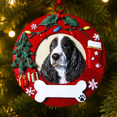 A round red Christmas ornament featuring a custom pet portrait of a black and white Springer Spaniel. The ornament is decorated with festive holiday elements including a green Christmas tree, red gifts, a red stocking, holly, snowflakes, and a white bone at the bottom, hanging on a Christmas tree with warm lights in the background.