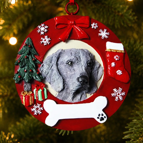 A circular red Christmas ornament hanging on a Christmas tree, featuring a portrait of a Weimaraner  in the center, surrounded by a red wreath-style border with a red bow, white snowflakes, a green Christmas tree, wrapped gifts, a red stocking, a white bone, and small white paw-print details. The ornament is photographed against blurred green pine branches with warm lights.