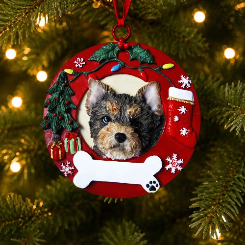 A round red Christmas ornament hanging on a Christmas tree, featuring a photo cutout of a Yorkshire Terrier's face surrounded by festive decorations including a green tree, white bone, snowflakes, and a red stocking.