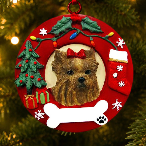 A red Christmas wreath-shaped ornament featuring a portrait of a Yorkshire Terrier dog in the center, surrounded by holiday decorations including a small Christmas tree, ornaments, a stocking, snowflakes, a bone shape, and a red hanging loop. The ornament is photographed hanging on a Christmas tree with warm blurred lights in the background.