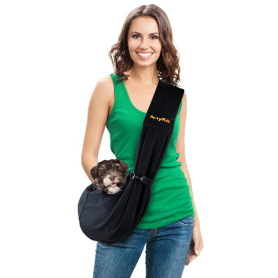 A woman wearing a green tank top and blue jeans carries a small brown dog in a black pet sling carrier with a wide shoulder strap. The brand name 'Furry Fido' is visible in yellow on the strap.