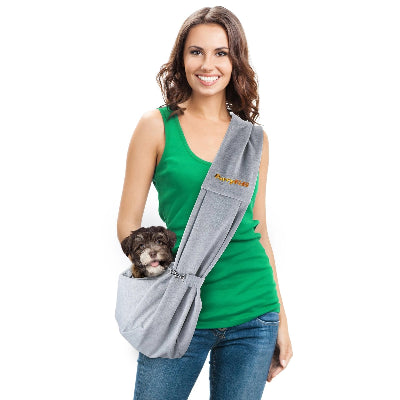 A woman wearing a green tank top and blue jeans models a gray pet carrier sling with a small brown dog peeking out from the front pocket. The sling has a small gold logo on the shoulder strap.