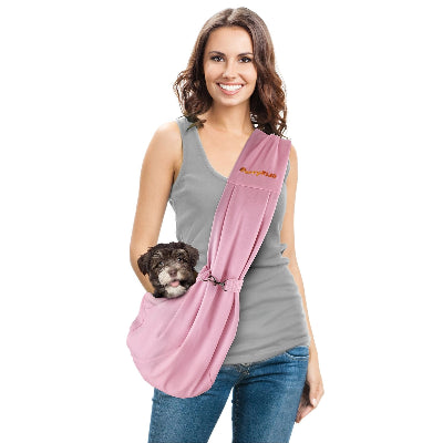 A woman wearing a gray tank top and blue jeans models a pink pet carrier sling with a small dark puppy inside, set against a plain white background.