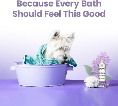 A small white dog sits in a lavender bath tub wrapped in a teal towel, with the text 'Because Every Bath Should Feel This Good' above and a bottle of pet shampoo, a brush, and purple flowers on a lavender surface below.
