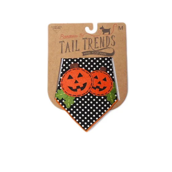 A Halloween-themed bandana for dogs, featuring two orange jack-o'-lantern faces on a black and white polka dot background with green leaves, presented on its original brown cardboard backing.